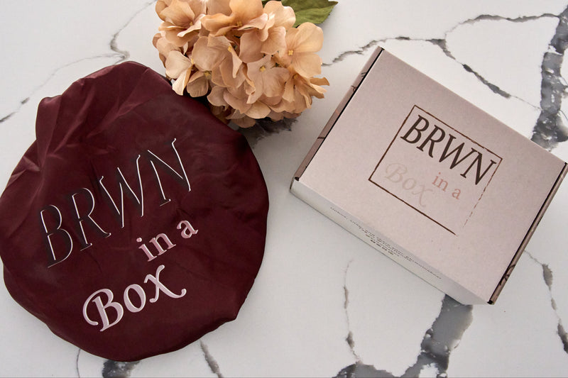 Brown satin bonnet, with a box 'BRWN (Brown) in a Box' branding on a marble surface with a flower